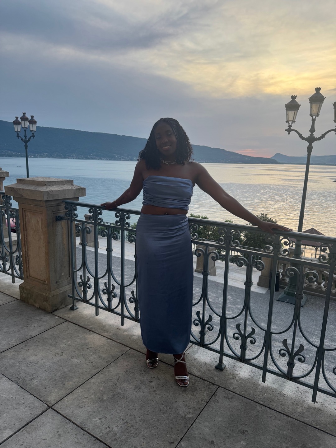 Rashana beside Lake Annecy at sunset with mountains and ornate lamps behind her